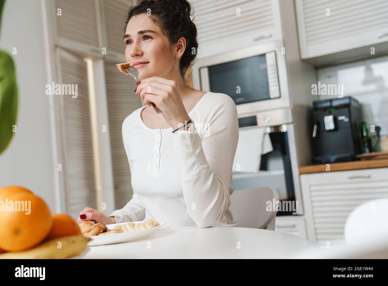 La femme souriante tenant un morceau de pain près de sa bouche tout en étant assise à la table dans une cuisine lumineuse Banque D'Images