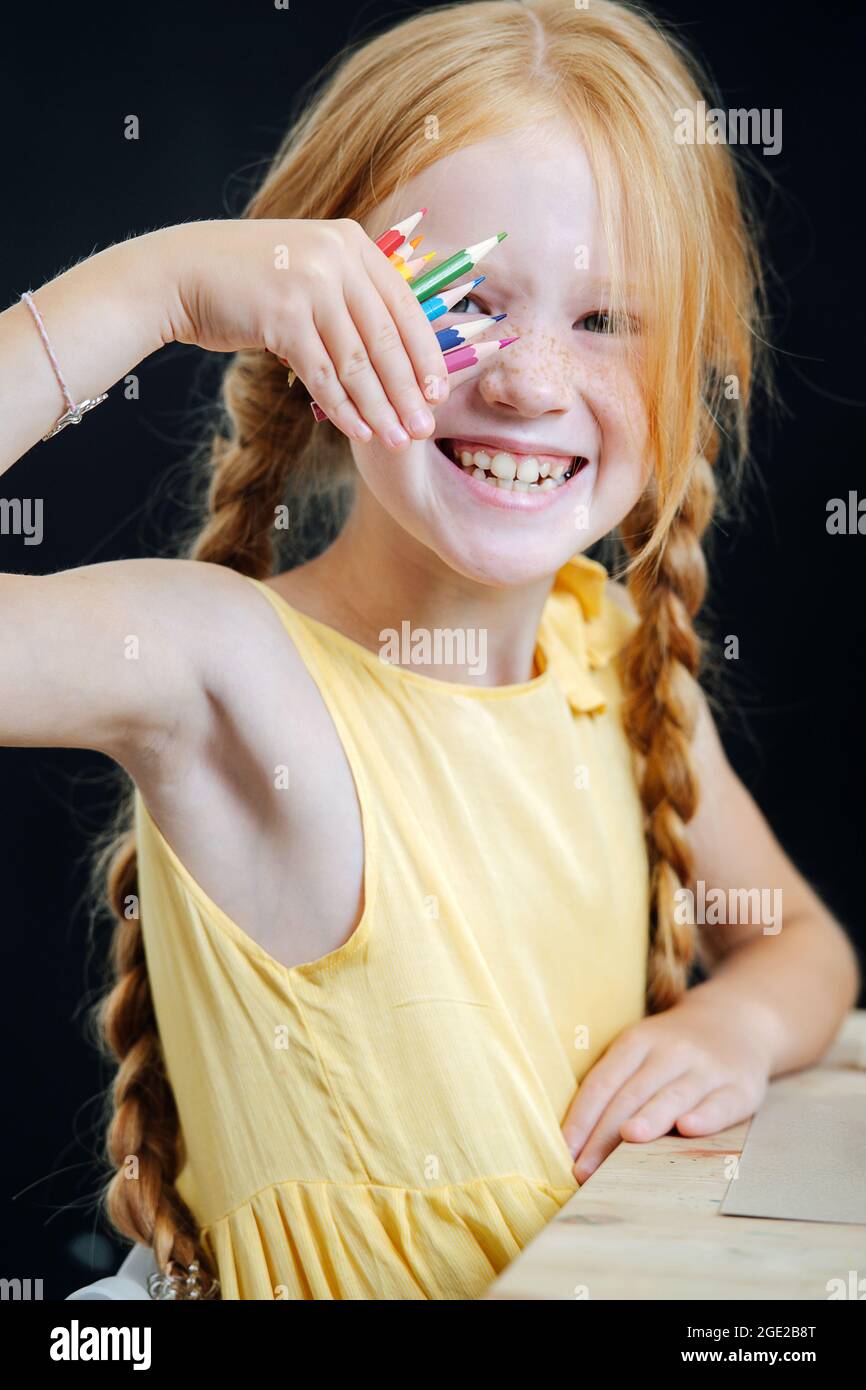 Ginger fille de sourire avec de longues tresses assis derrière une table, montrant des crayons Banque D'Images