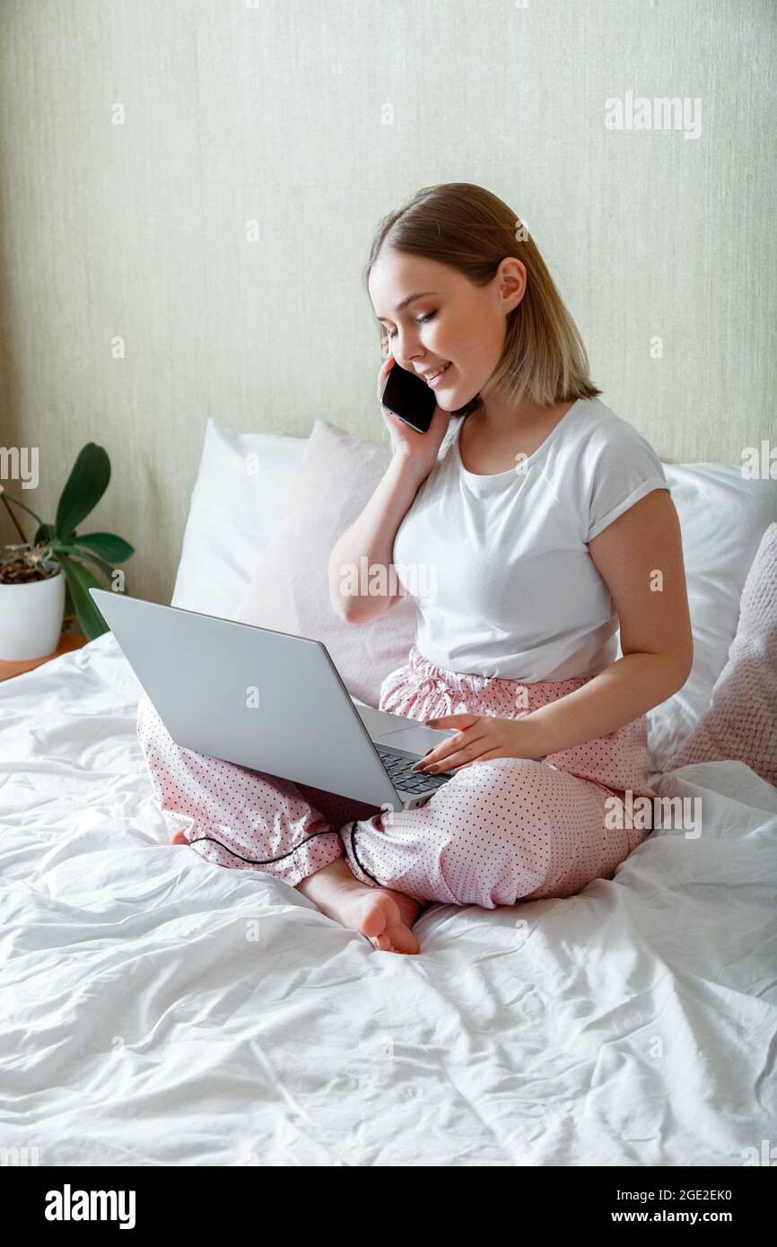 Jeune femme souriante en pyjama assis dans le lit de travail utiliser un ordinateur portable smartphone le matin. Conversation professionnelle par téléphone le matin. Femme d'affaires Banque D'Images