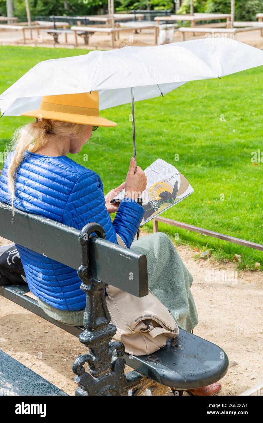 Dame dans un manteau bleu, chapeau jaune et parapluie blanc lisant sur un banc dans les Jardins du Luxembourg à Paris Banque D'Images