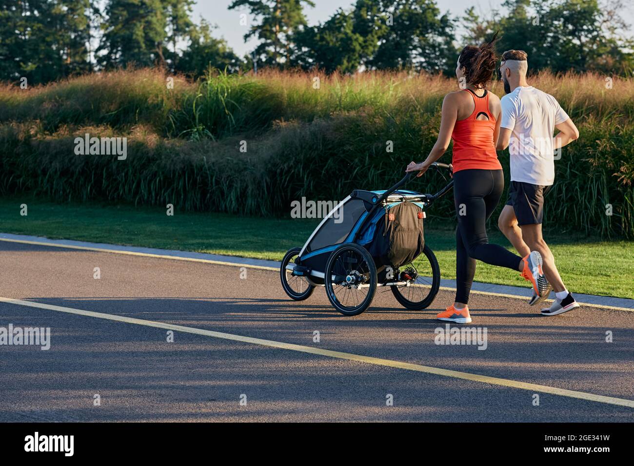 courir jeune famille avec poussette de jogging bébé en mouvement profiter de l'entraînement et la journée d'été dans le parc de la ville. Vue arrière Banque D'Images