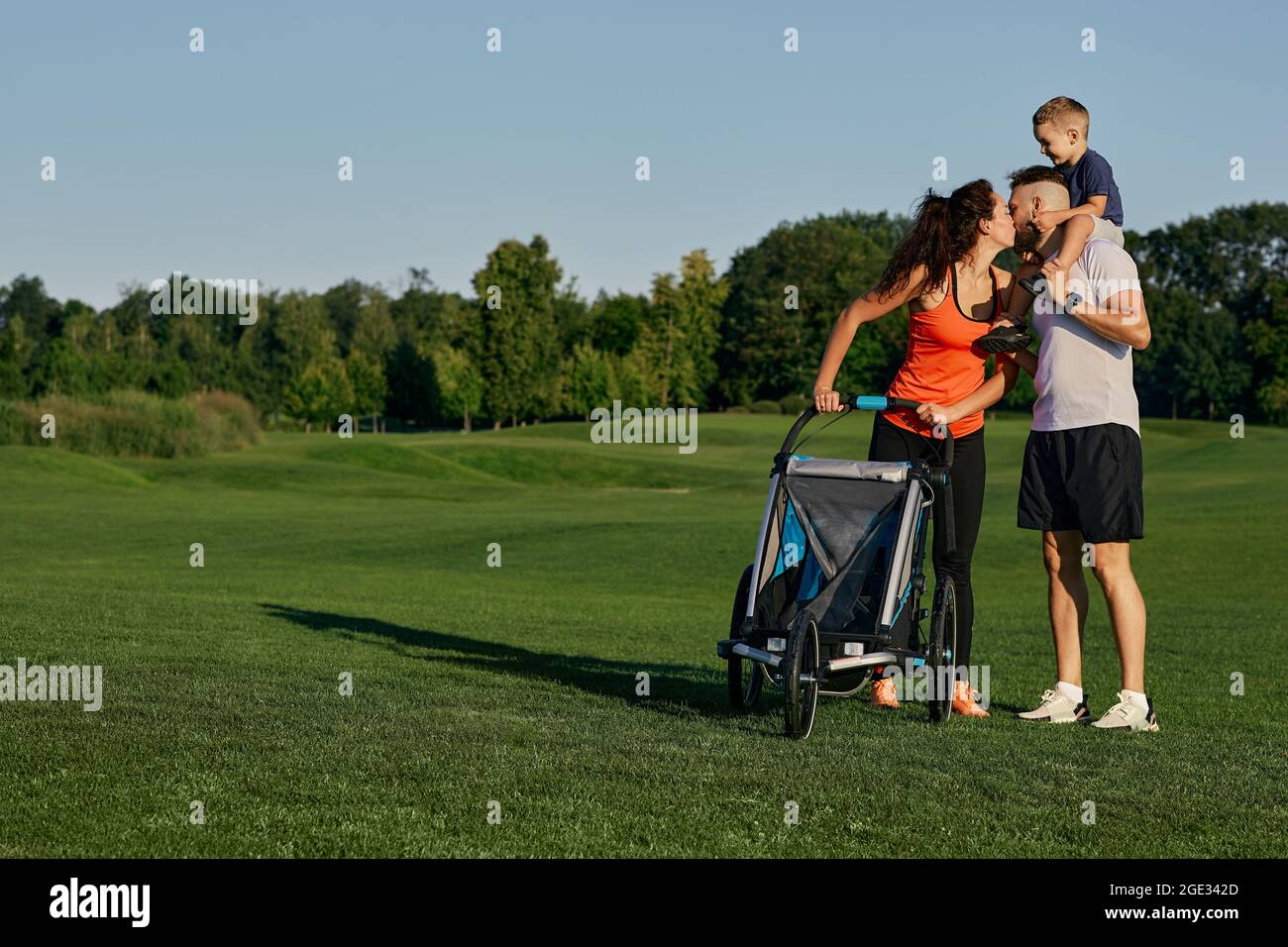 Une jeune famille heureuse avec un enfant passe du temps ensemble tout en marchant avec une poussette sur une pelouse verte dans un parc de la ville pendant un week-end. Jour de la famille Banque D'Images