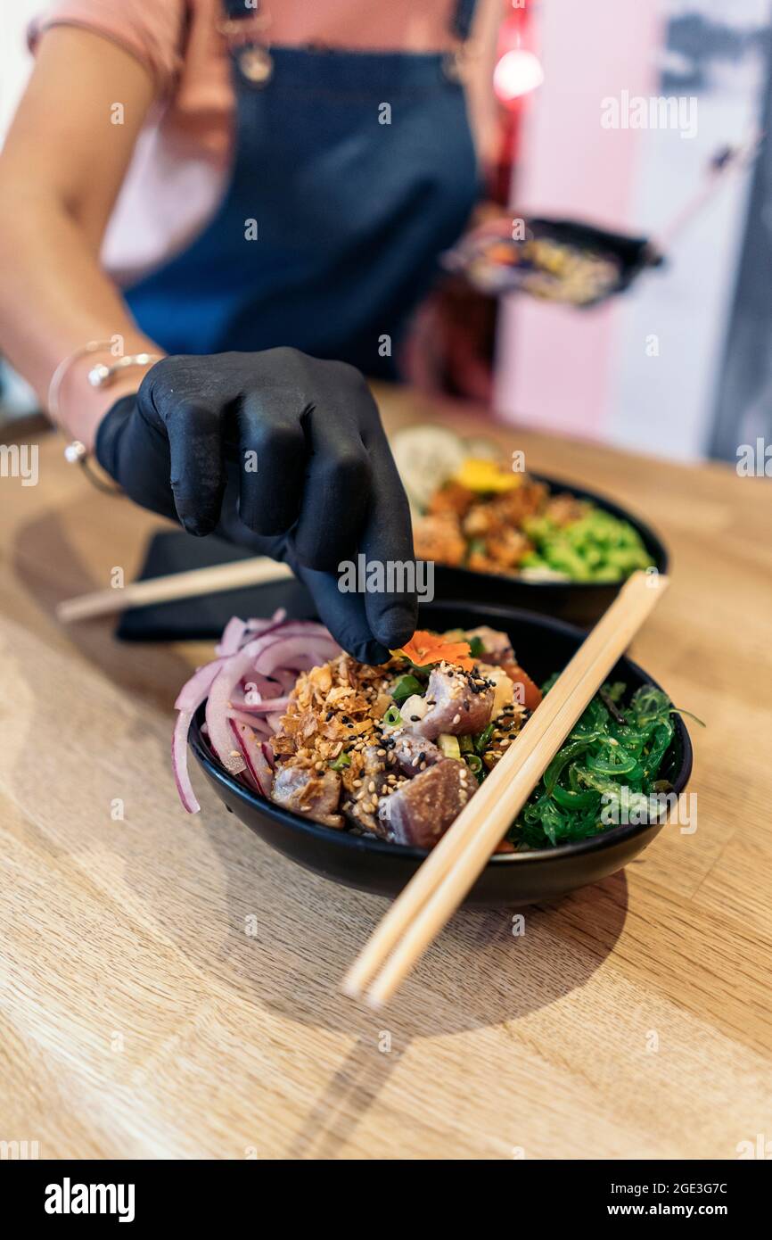 Employé de restaurant non reconnu dans des gants de protection mettant des ingrédients dans un bol de poke avec du riz et des légumes. Banque D'Images