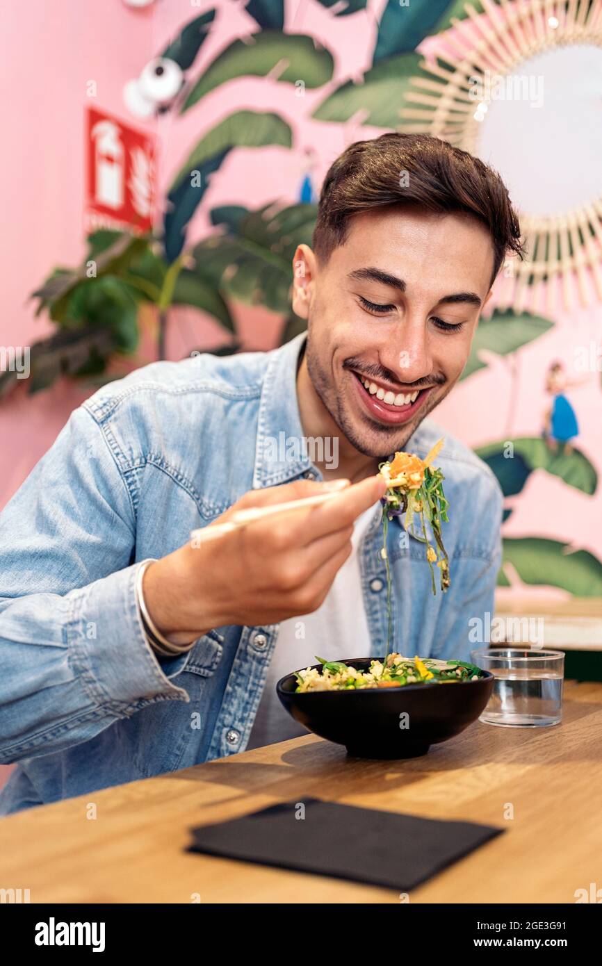 Jeune homme assis dans un restaurant en appréciant un poke-Bowl sain et en utilisant des baguettes. Banque D'Images