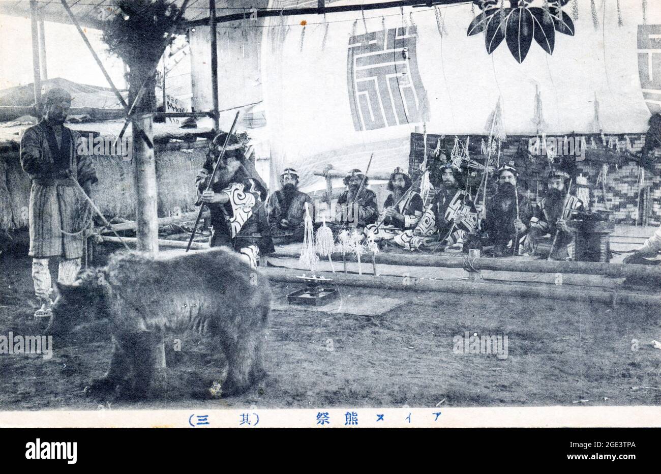 Ancienne carte postale japonaise noire et blanche, vers 1910, d'un groupe de personnes Ainu assis, portant un costume traditionnel, avec un ours captif attaché devant. Banque D'Images