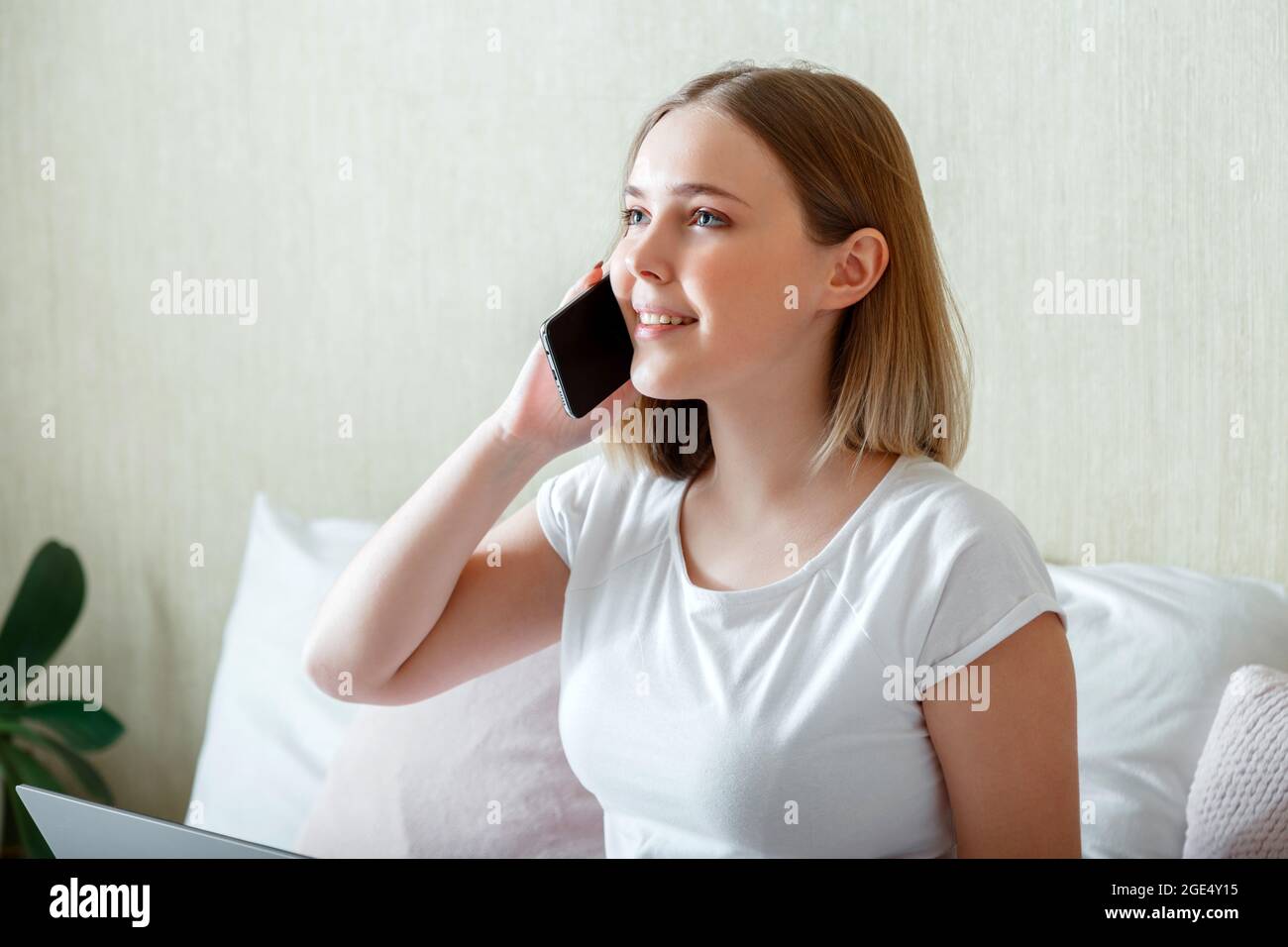 Jeune femme souriante parlant par téléphone dans la chambre. Conversation avec un téléphone intelligent le matin à l'intérieur de la maison. Bonne jeune femme blonde de race blanche communiquer Banque D'Images