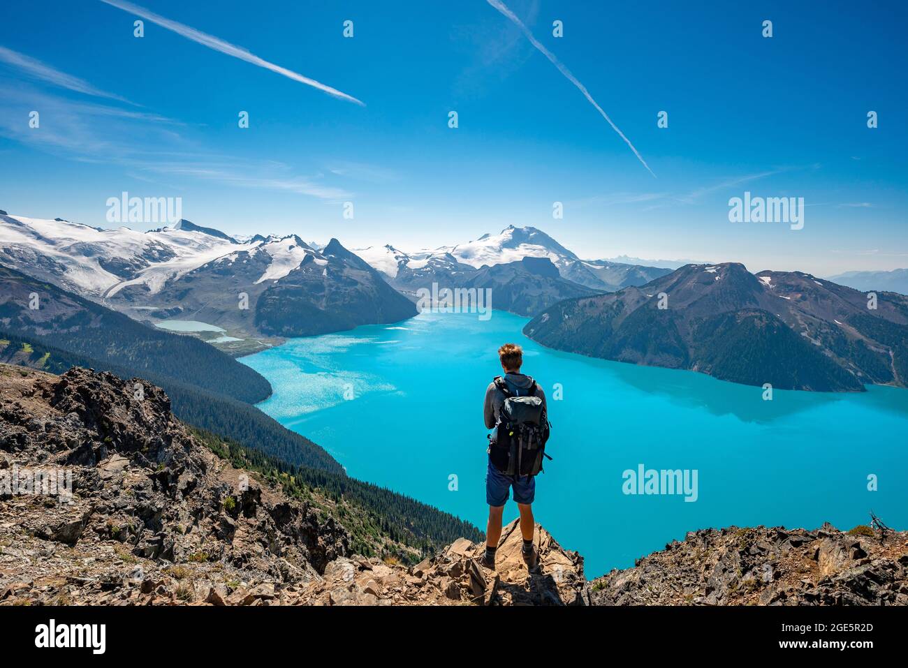 Jeune homme debout sur un rocher, regardant au loin, vue sur les montagnes et le glacier avec le lac bleu turquoise Garibaldi Lake, Peaks Panorama Ridge Banque D'Images