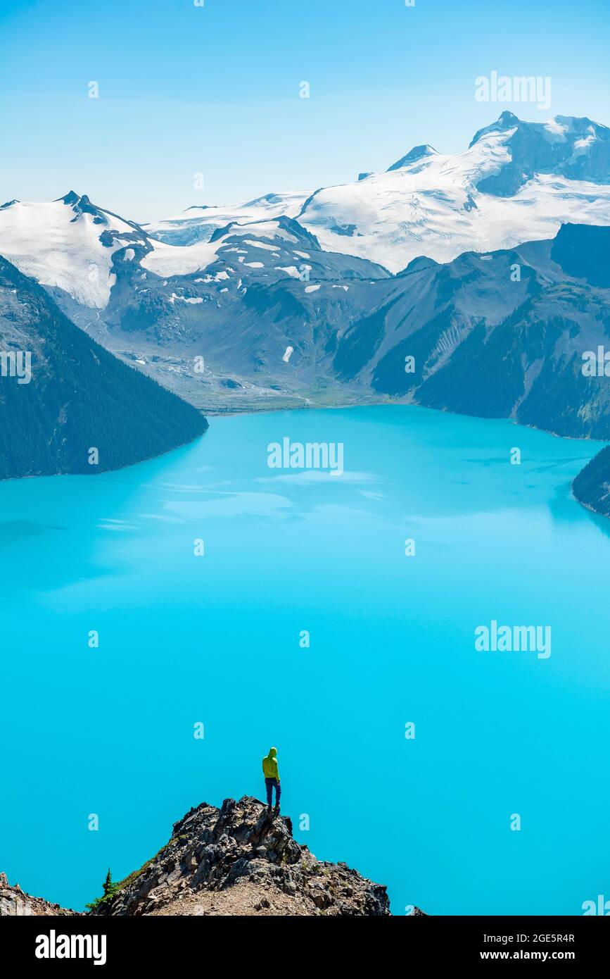 Jeune homme debout sur un rocher, regardant au loin, vue sur les montagnes et le glacier avec le lac bleu turquoise Garibaldi Lake, Peaks Panorama Ridge Banque D'Images