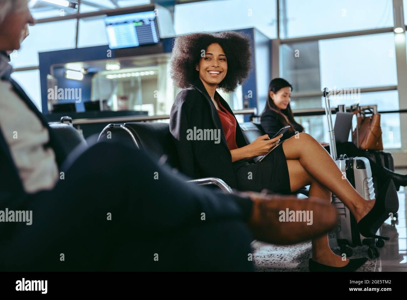 Bonne femme aux cheveux bouclés assise à l'aéroport. Femme d'affaires attendant au terminal de l'aéroport avec des gens assis autour. Banque D'Images