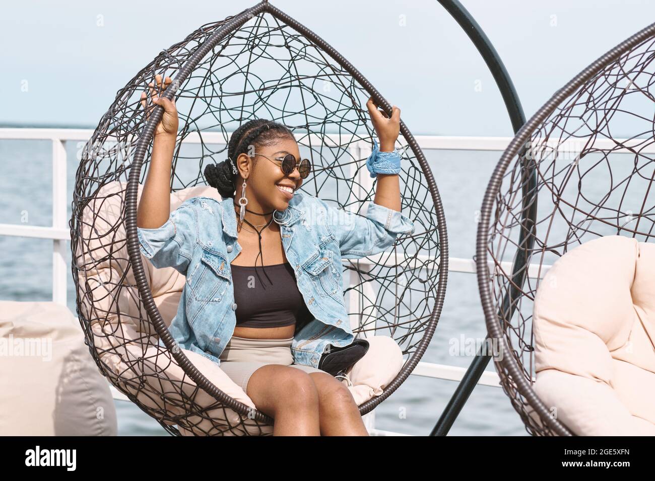 Joyeuse et élégante jeune femme en veste en denim, lunettes de soleil et longues boucles d'oreilles assis dans une chaise suspendue lors d'une fête d'anniversaire en plein air Banque D'Images