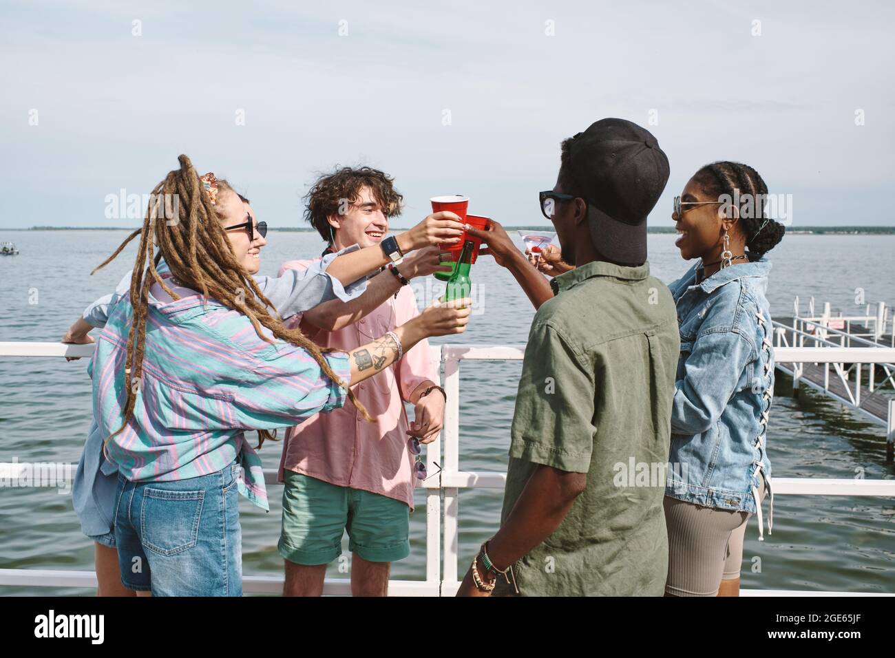 Joyeux jeunes gens joyeux qui toaster avec des verres et des bouteilles de bière lors d'une petite fête sur la jetée Banque D'Images