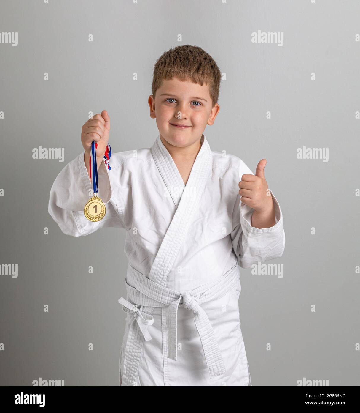 Premier prix. Garçon dans un kimono blanc avec des meubles en or à la main. Un enfant satisfait se montre un pouce vers le haut. Arrière-plan gris. Banque D'Images