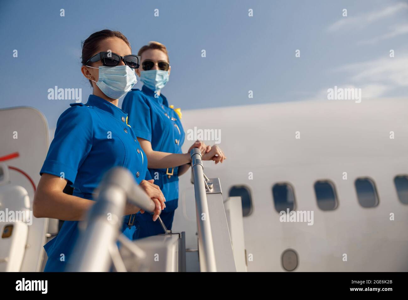 Deux jeunes hôtesses d'air en uniforme bleu, des lunettes de soleil et des masques de protection qui regardent la caméra, debout sur un escalier d'air pendant la journée Banque D'Images