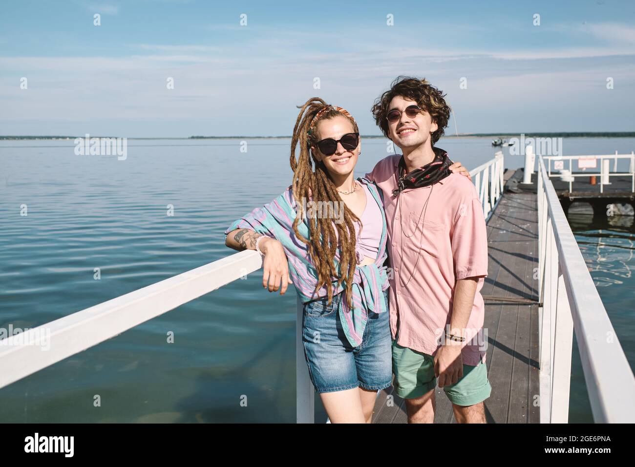 Envieux gai jeune petit ami et petite amie debout sur la jetée, embrassant et regardant l'appareil photo Banque D'Images