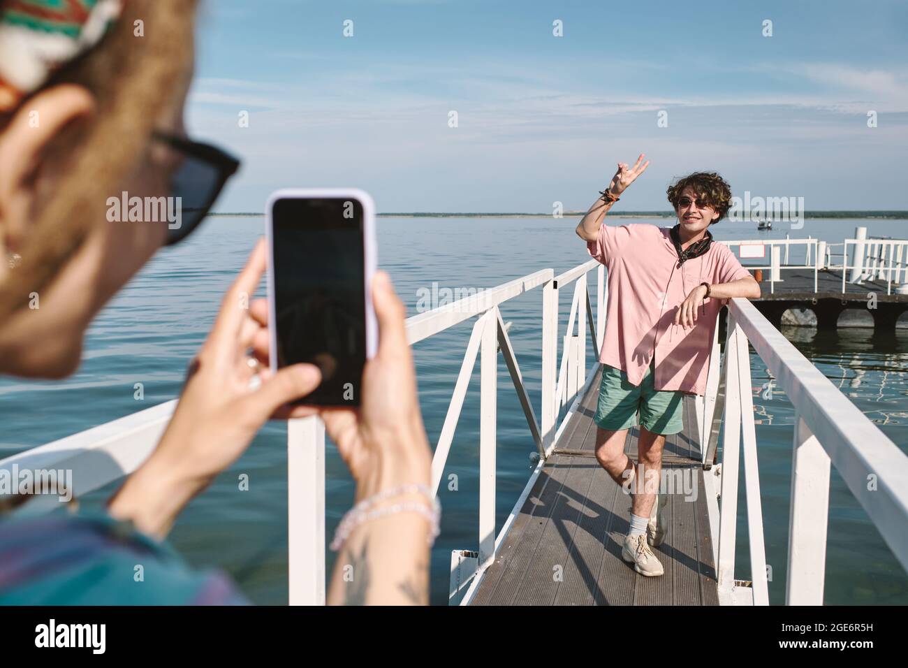 Un jeune homme souriant en chemise rose fait un geste de paix lorsque sa petite amie le photographie sur un smartphone au bord du lac Banque D'Images