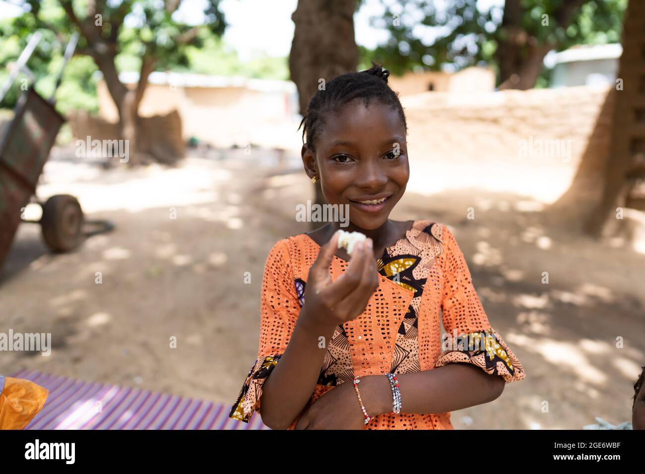 Élégante fille africaine noire debout dans la cour de sa famille, se grinçant à l'appareil photo tout en mangeant un œuf dur Banque D'Images