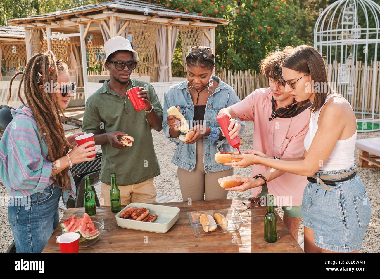 Divers groupes d'amis buvant des bières et mangeant des hot dogs à table sur la plage en profitant de la petite fête d'été Banque D'Images