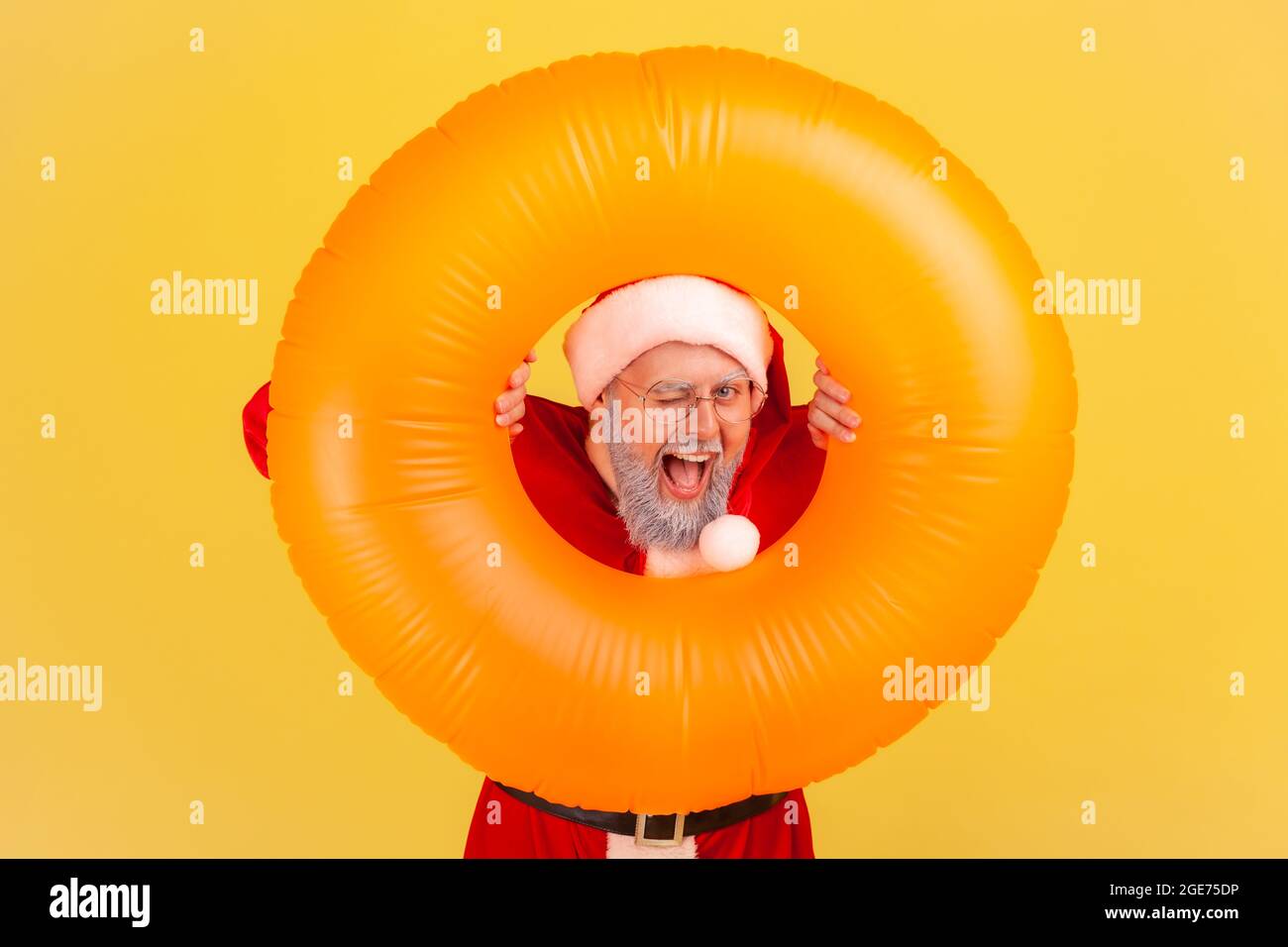 Homme âgé satisfait avec une barbe grise portant le costume du père noël tenant l'anneau en caoutchouc entre les mains, regardant l'appareil photo et le wencing, visite de Noël. Intérieur Banque D'Images