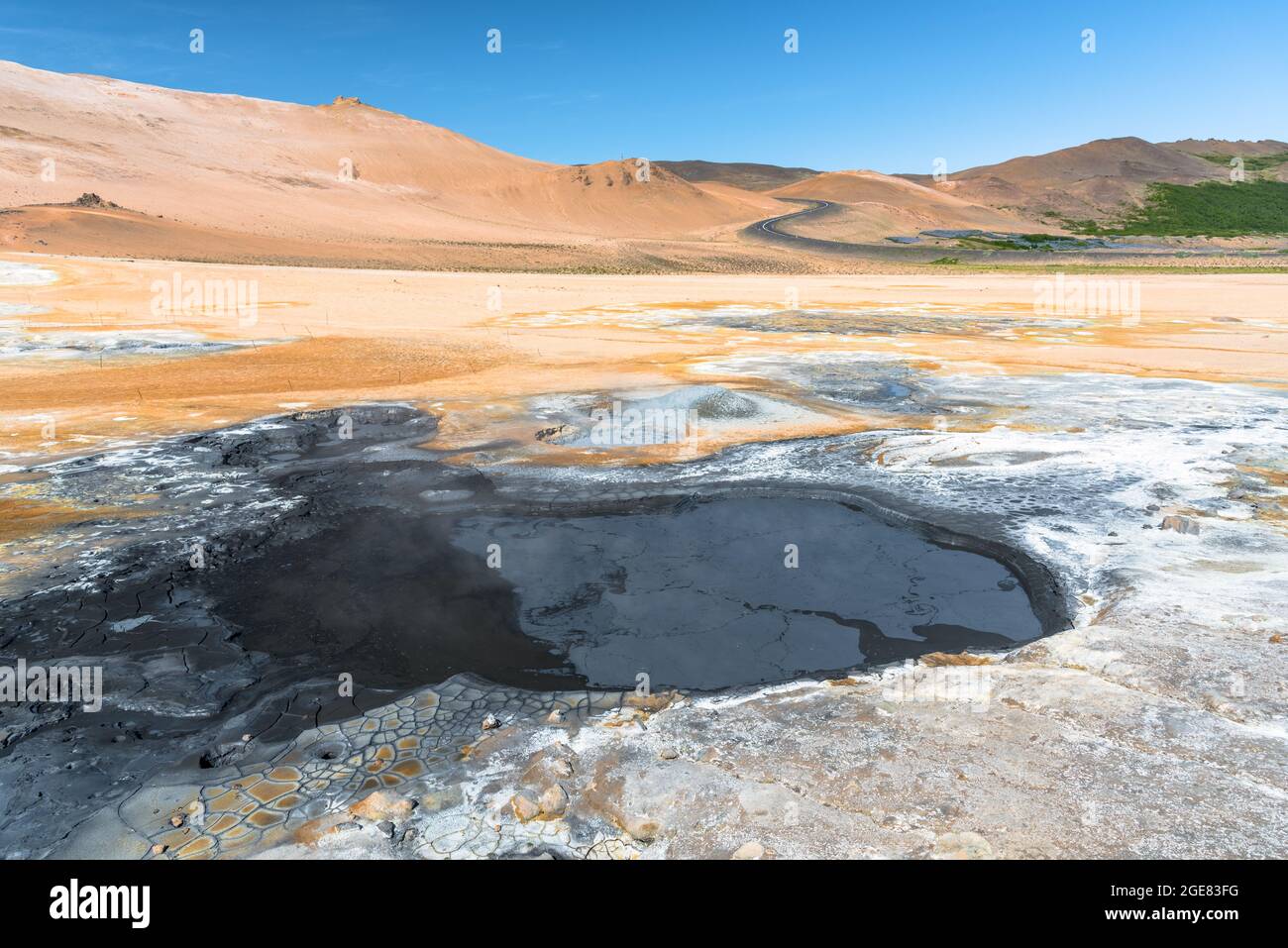 Piscine de boue bouillonnante dans une zone géothermique en Islande, par une belle journée d'été. Une route sinueuse grimpant une colline volcanique aride est visible en arrière-plan. Banque D'Images