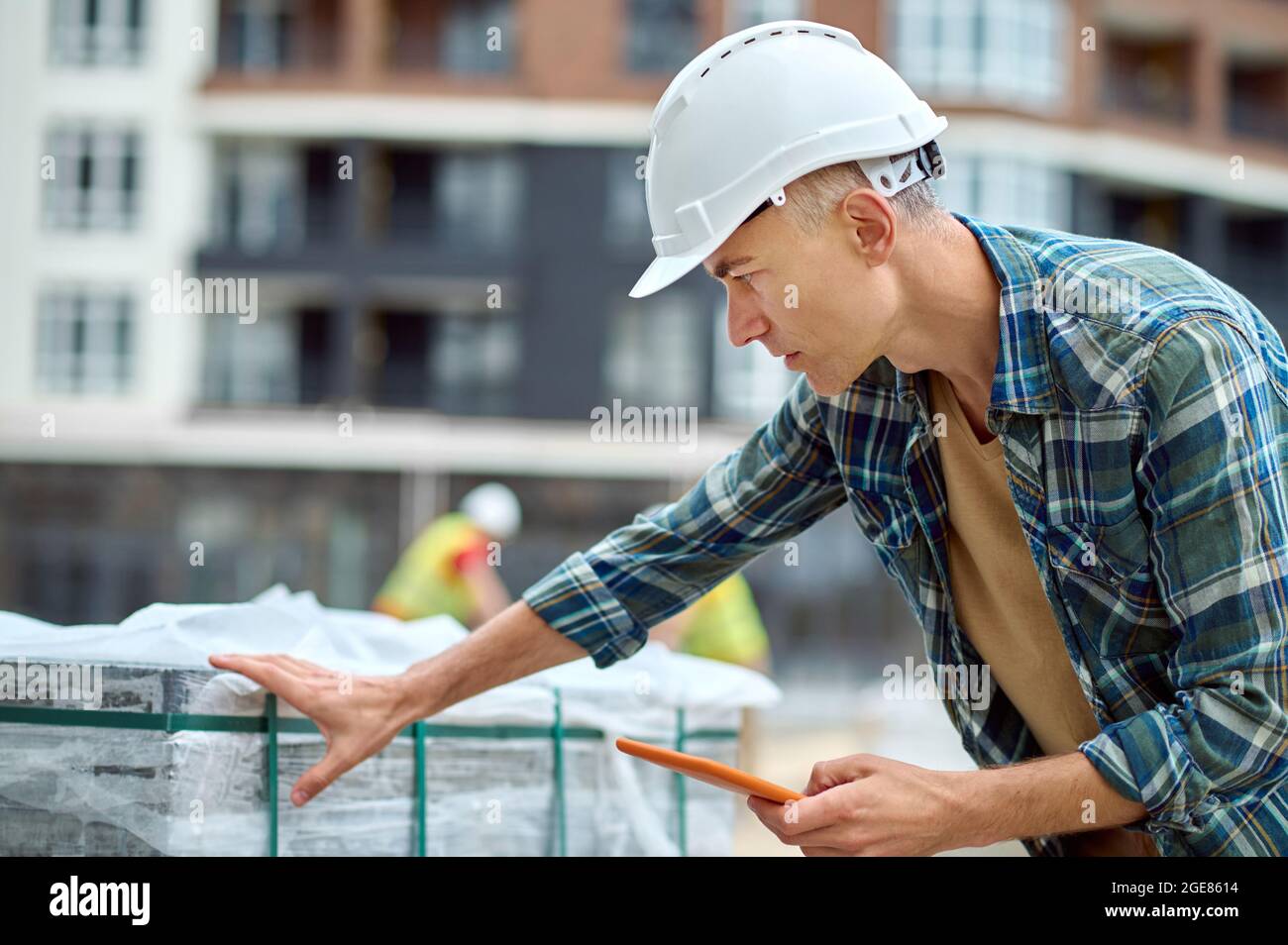 Travailleur concentré sérieux dans un casque de chantier vérifiant les matériaux de construction à l'extérieur Banque D'Images
