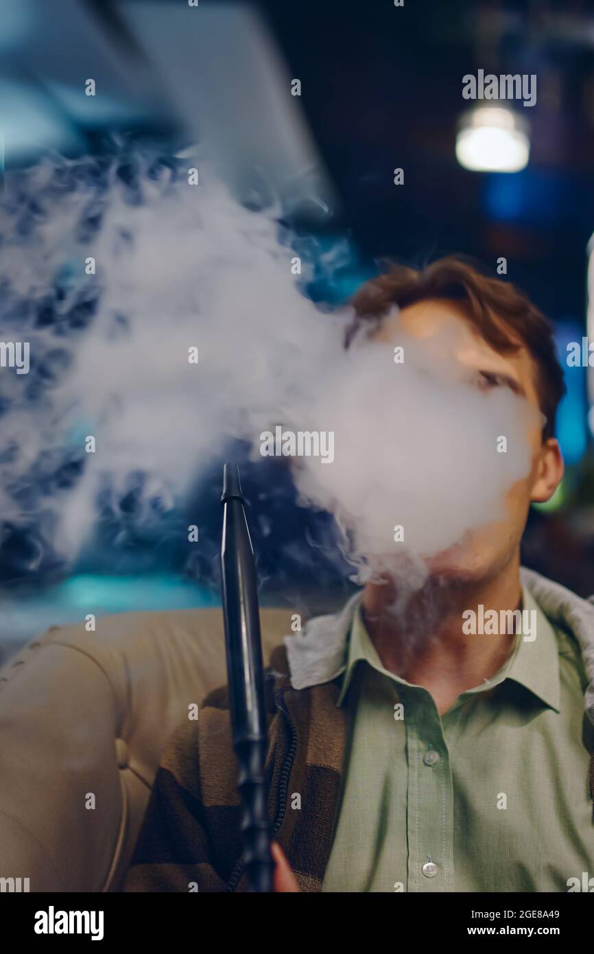 Homme dans le café fumant le narguilé. Fumée épaisse de shisha. Jeune homme sur le canapé dans le restaurant. Des lumières de couleur s'allument en arrière-plan. Détendez-vous au bar shisha. Banque D'Images