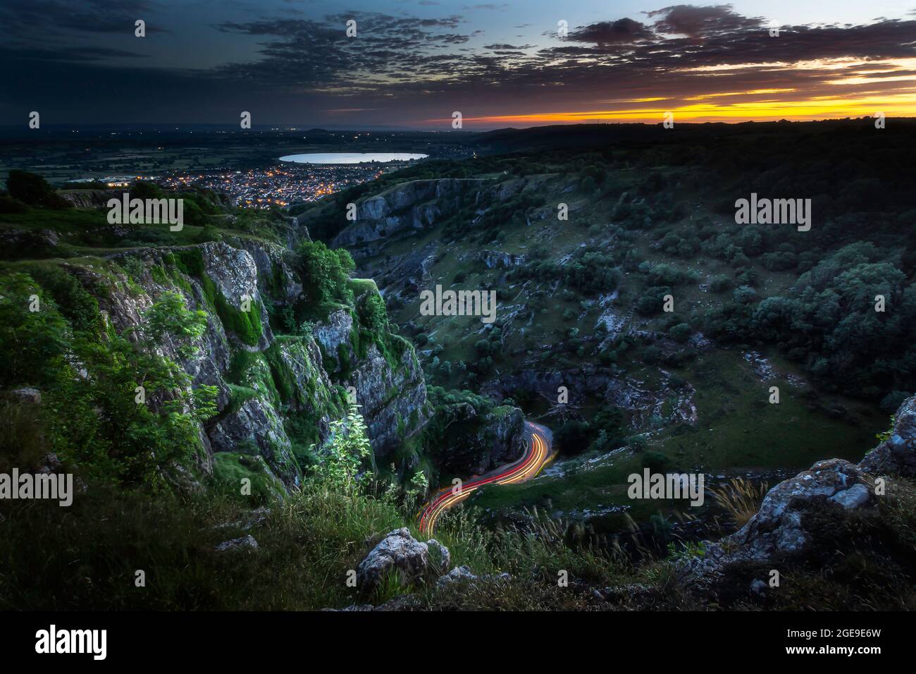 Soirée dans la gorge de Cheddar, Somerset, Royaume-Uni. Paysage de l'heure bleue avec une lueur orange sur ciel nuageux, essais de lumière de voiture et lumières de ville en arrière-plan Banque D'Images