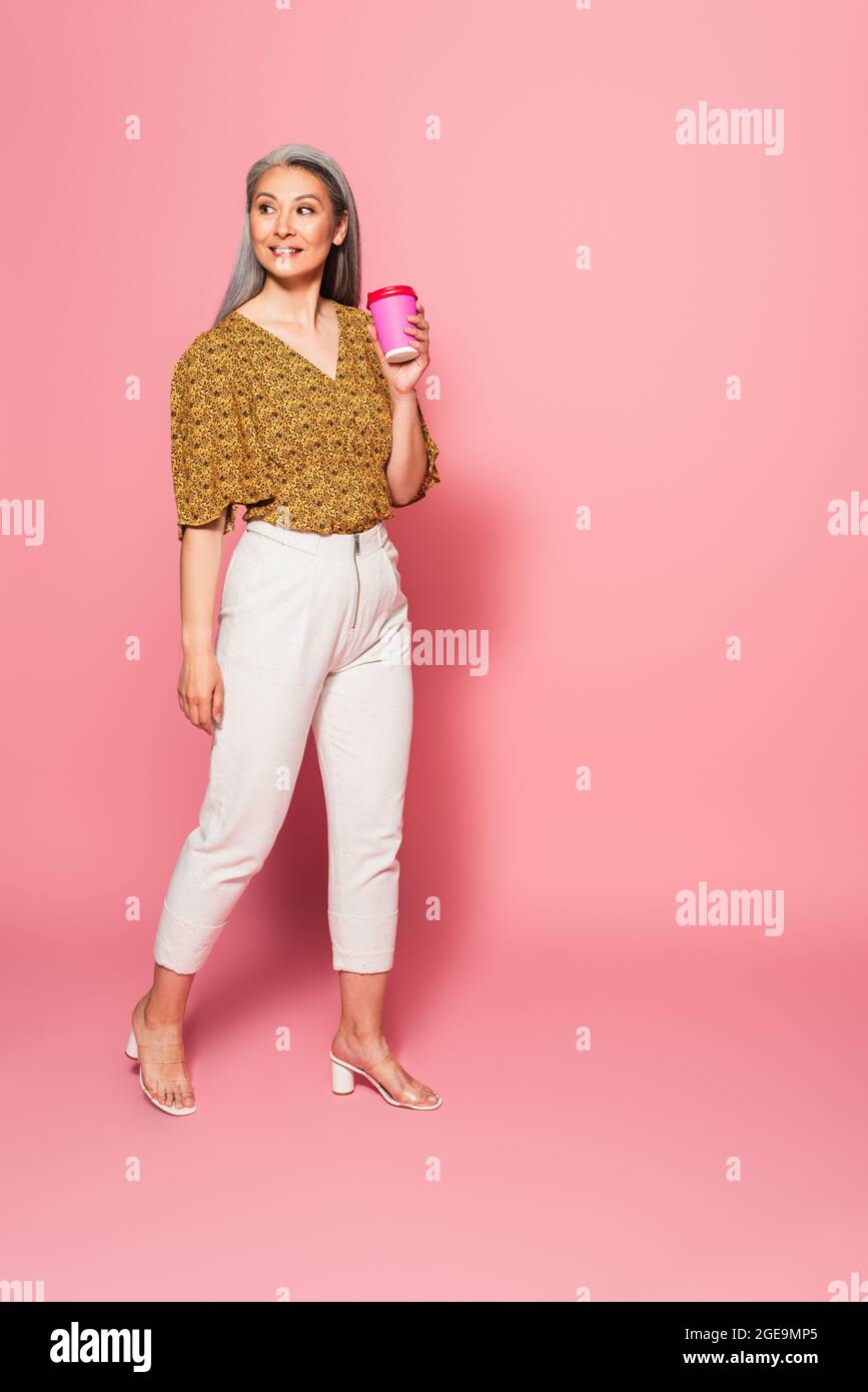 femme adulte dans un chemisier à motifs et pantalon blanc marchant avec une boisson à emporter sur du rose Banque D'Images