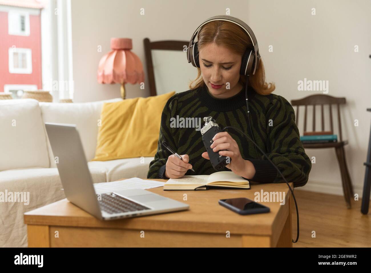 Animateur de radio féminin focalisé avec micro et casque écrivant dans le bloc-notes tout en préparant l'enregistrement de podcast à la maison Banque D'Images