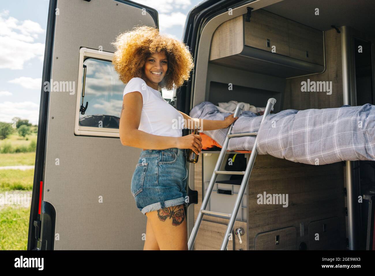 Vue latérale d'une femme noire ravie avec de la bière riant et regardant la caméra tout en se tenant sur l'échelle à l'extérieur de la campeuse et en montant sur le lit le jour d'été Banque D'Images