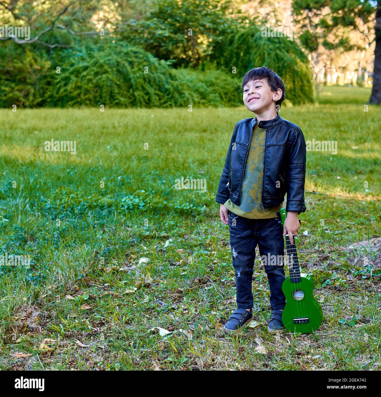 latino garçon debout souriant dans la forêt tenant un ukulele vert à ses côtés. carré Banque D'Images