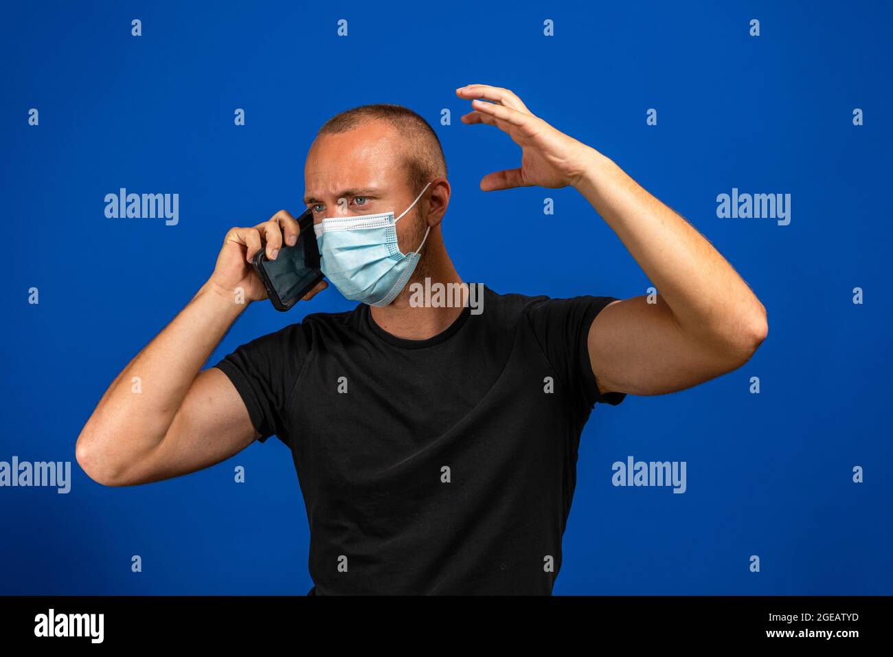Jeune caucasien parlant sur le téléphone mobile très en colère, il porte un masque chirurgical pour se protéger du coronavirus. Isolé sur studio bleu Banque D'Images