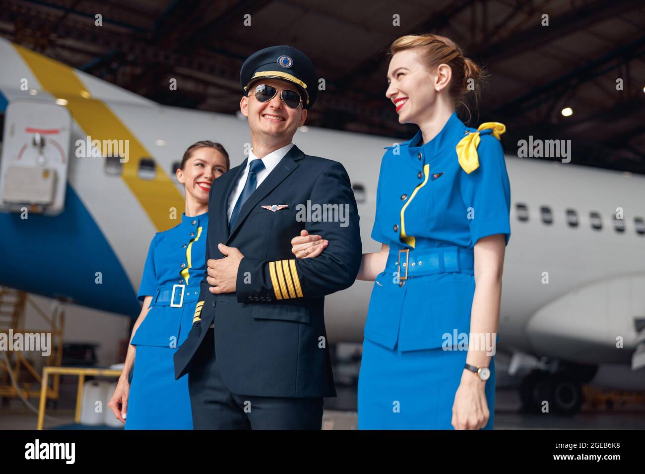 Pilote souriant en uniforme et lunettes de soleil aviateur marchant avec  deux hôtesses d'air en uniforme bleu devant un gros avion passager Photo  Stock - Alamy