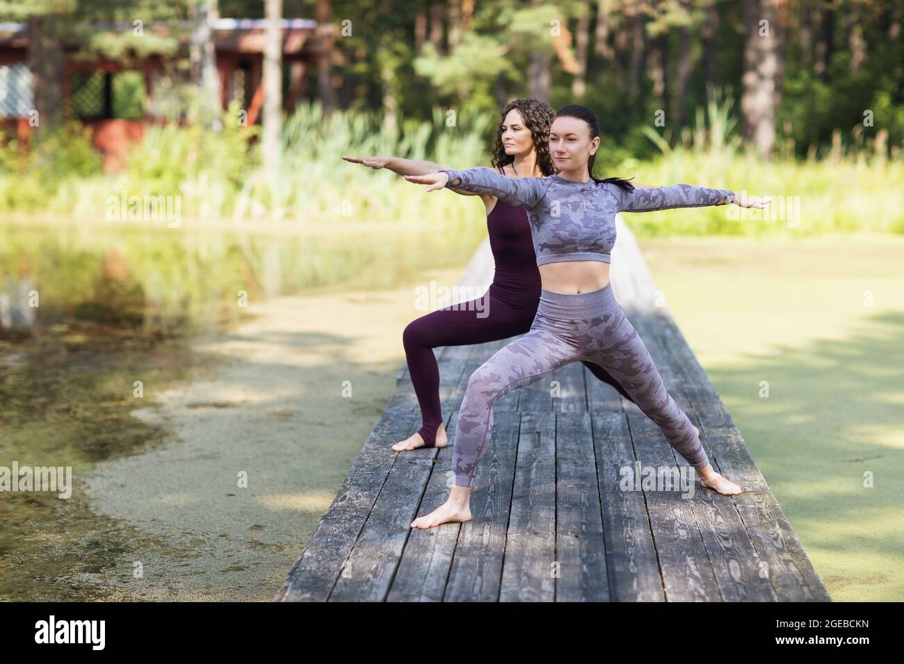 Deux femmes pratiquant le yoga le matin sur un pont en bois dans le parc exécutent l'exercice de virabhadrasana, posture de guerrier Banque D'Images