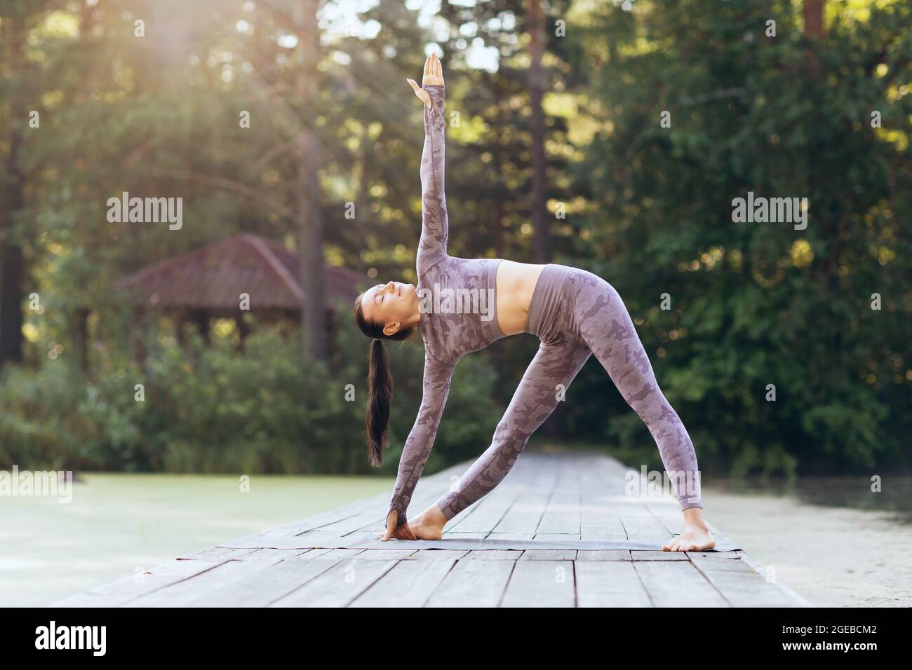 Jeune femme pratiquant le yoga, debout sur un tapis dans le parc un matin ensoleillé, faisant l'exercice uthita trikonasana, posture triangulaire Banque D'Images