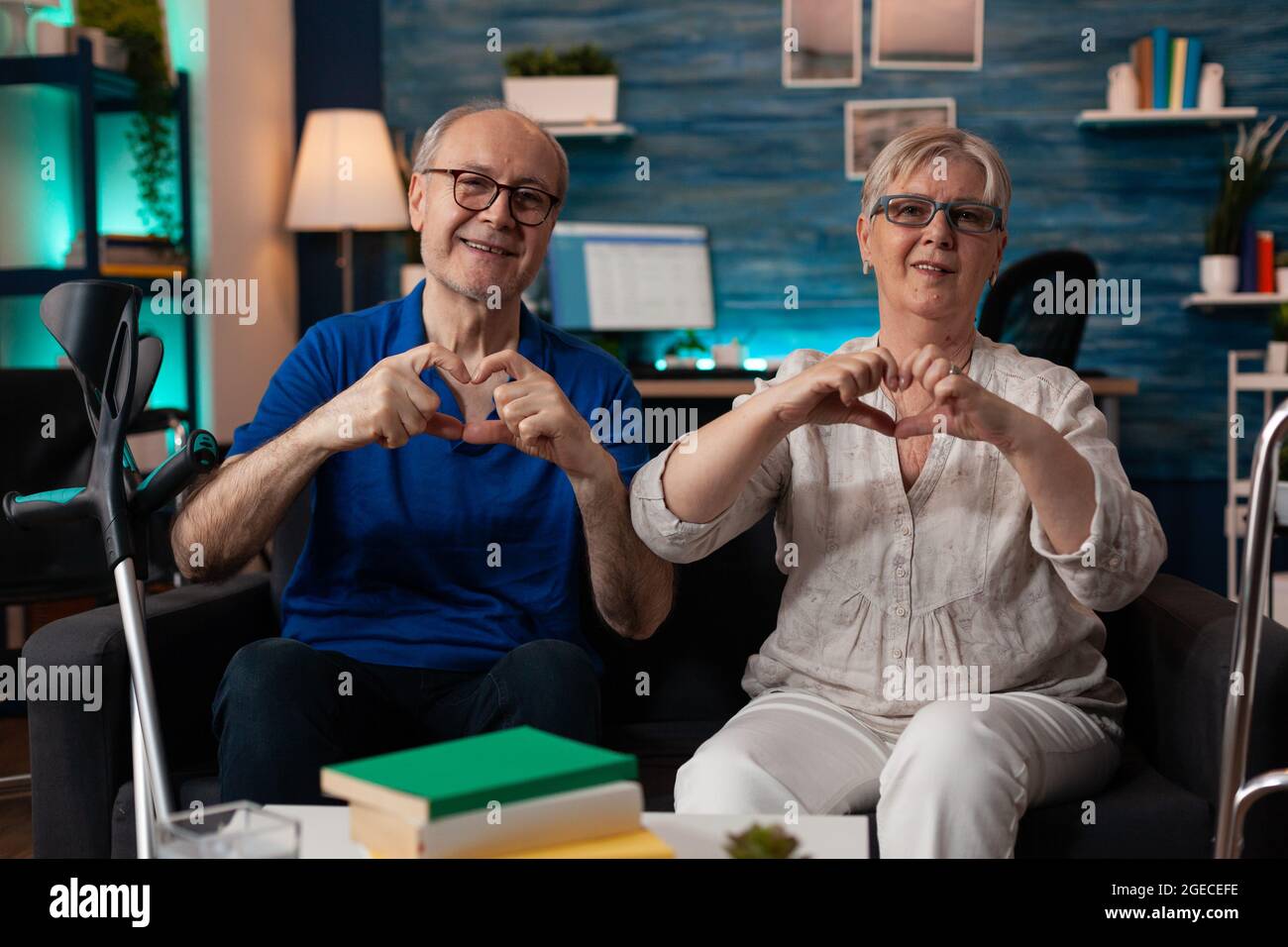 Vieux caucasien heureux couple faisant coeur signe avec les mains tout en regardant la caméra gaie assis dans la salle de séjour à la maison. Homme et femme gaie à la retraite montrant le symbole de l'amour romantique Banque D'Images