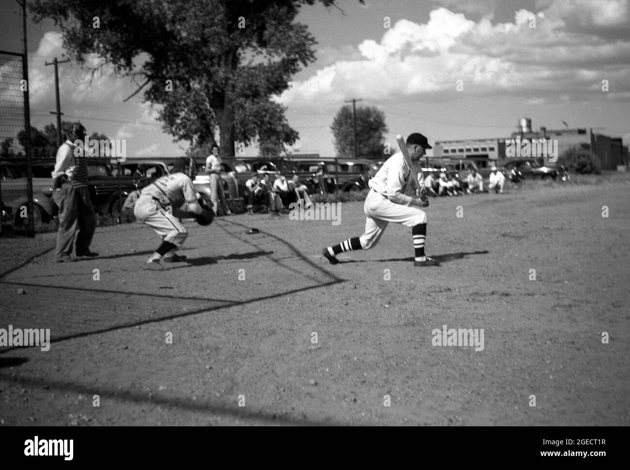 Années 1950, historique, à l'extérieur d'une zone poussiéreuse de terrain ouvert ou de terrain de balle, à côté d'une usine et avec des voitures de l'époque garées, les ouvriers de la construction locale en tenue complète jouant un match organisé et compétitif de baseball, Mid-West, USA. Ici, nous voyons un bavardage, un attrape et un juge-arbitre. Banque D'Images