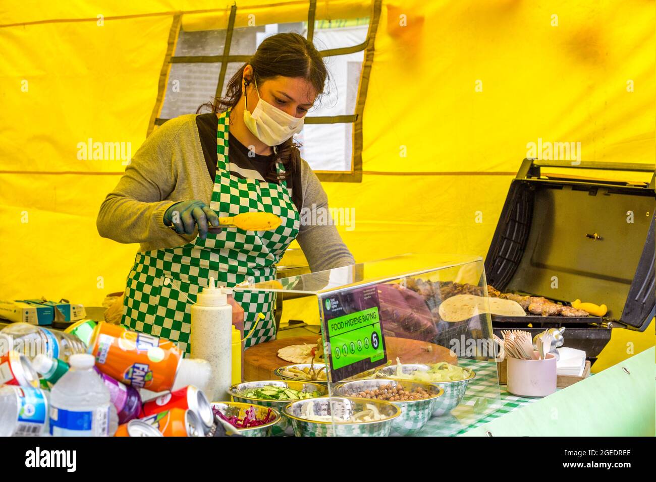 Une femme portant une couverture faciale, un tablier et des gants prépare de savoureux plats grillés de rue. Banque D'Images