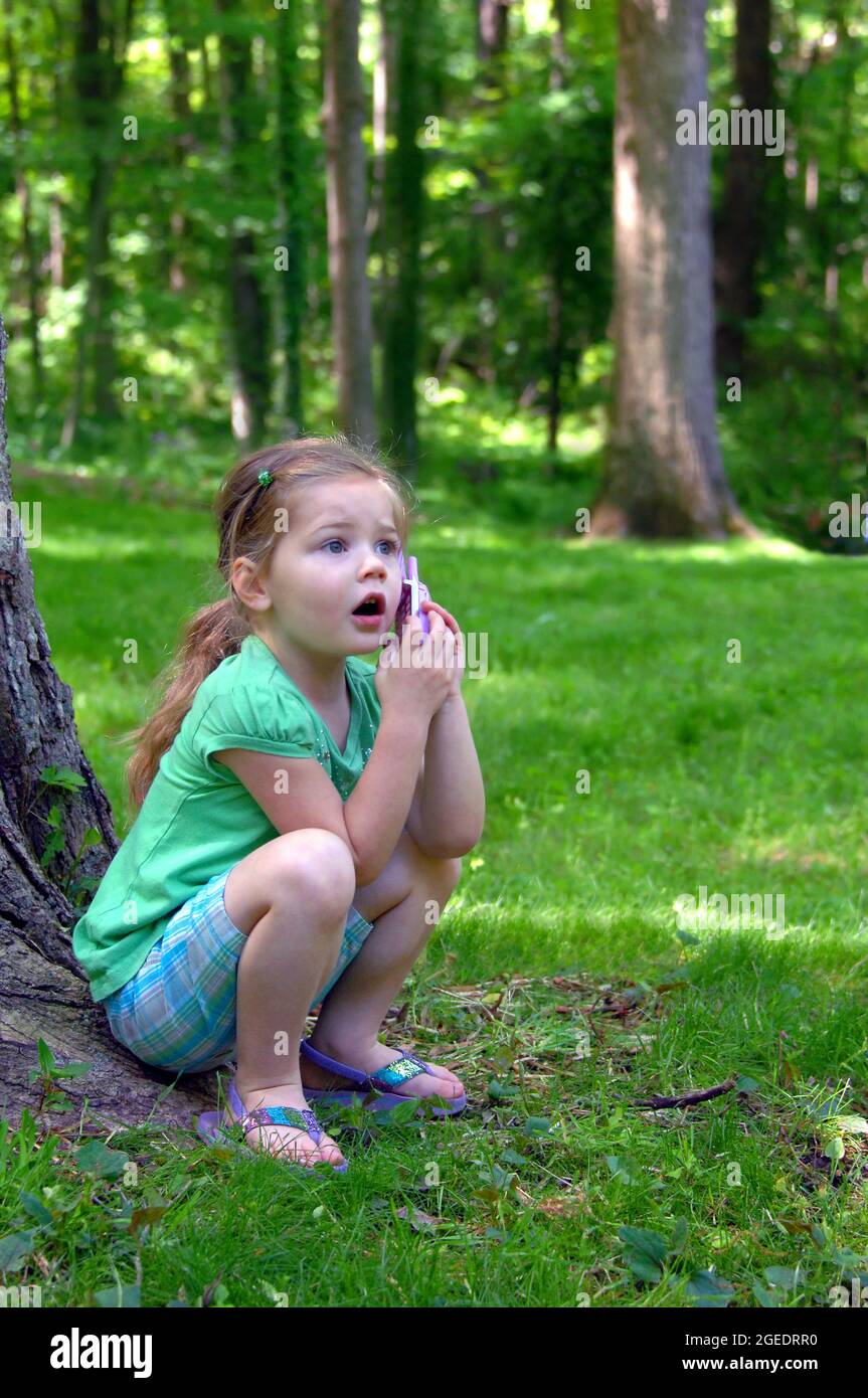 Petite fille montre son étonnement à reçu un vrai appel téléphonique. Elle joue à l'extérieur avec un téléphone à l'oreille. Elle est assise à côté d'un arbre. Banque D'Images