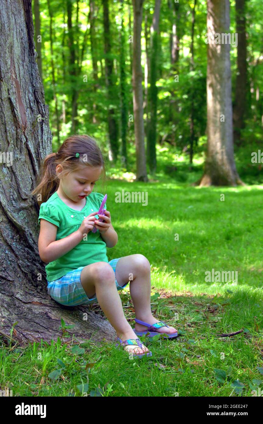 Une petite fille prétend appeler un ami sur son téléphone de jeu. Elle porte un short à carreaux et un haut vert. Elle est assise à côté d'un arbre sur le gr Banque D'Images