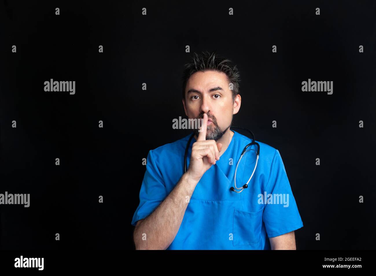 Un médecin vêtu d'un uniforme bleu d'hôpital, sombre et barbu, portant un stéthoscope autour de son cou, demande le silence avec son index Banque D'Images