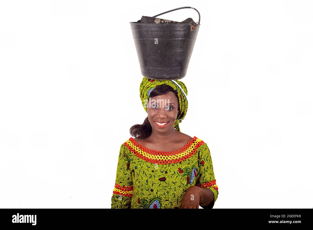 Belle femme adulte africaine vêtue de tissu de longe vert équilibre un seau noir sur sa tête et accompagnée d'un beau sourire Banque D'Images