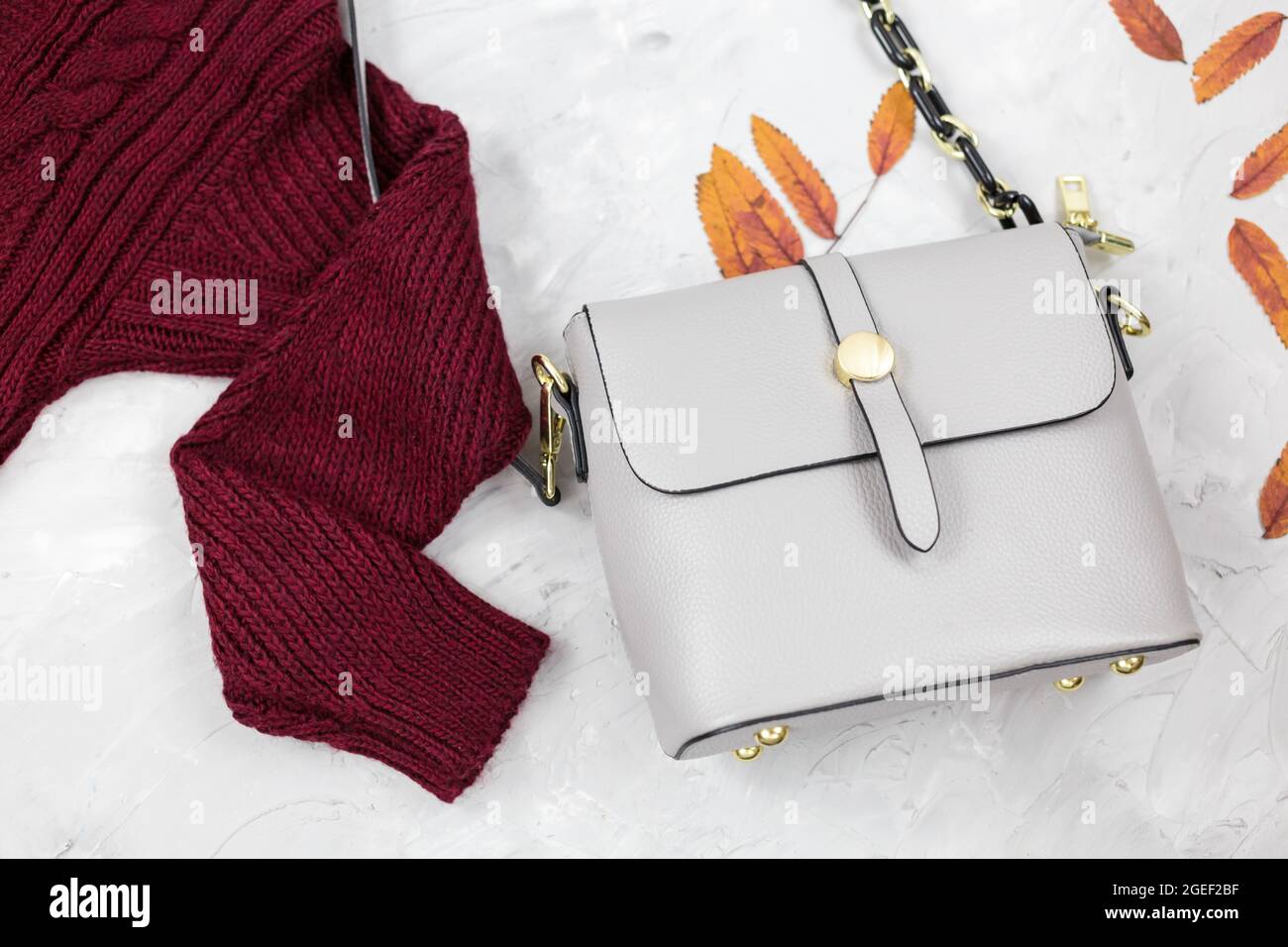 Sac femme en cuir gris, chandail rouge foncé, feuilles d'automne sur fond gris. Accessoires tendance pour l'automne. Tenue confortable Banque D'Images