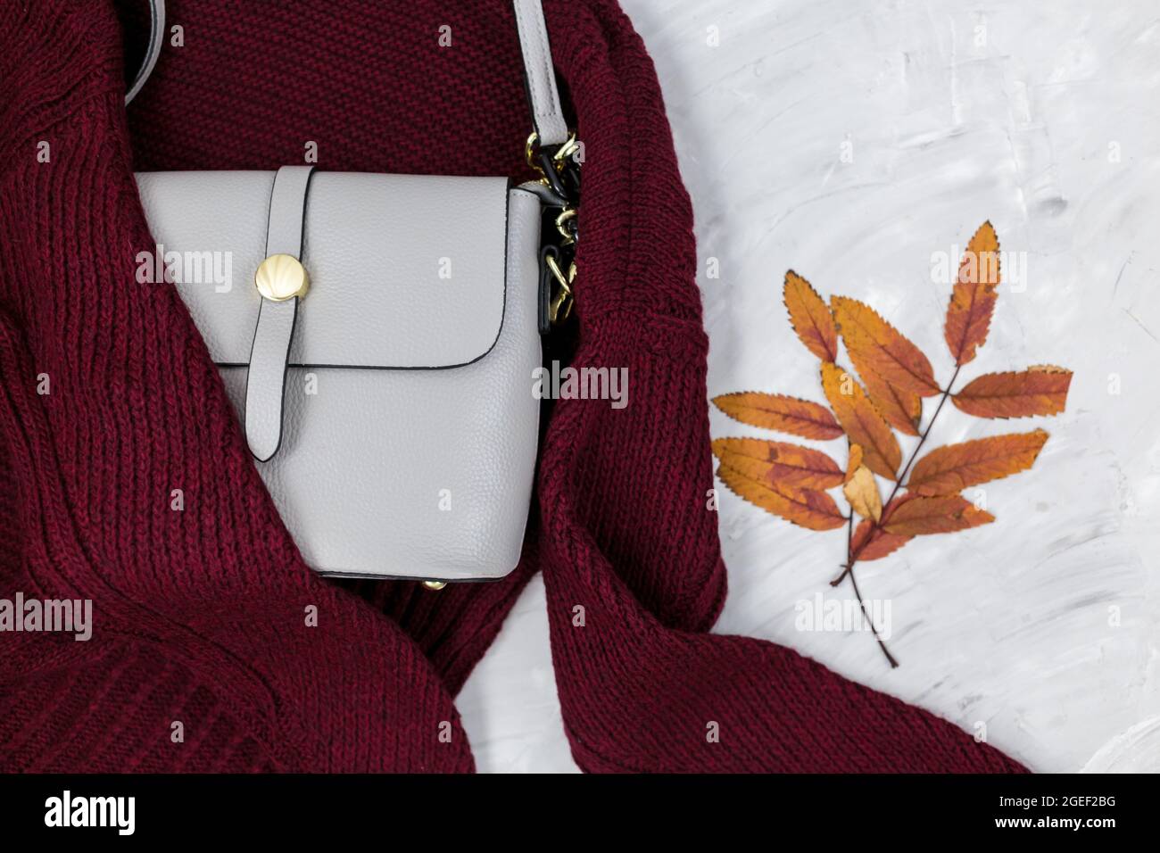 Sac femme en cuir gris, chandail rouge foncé, feuilles d'automne sur fond gris. Accessoires tendance pour l'automne. Tenue confortable Banque D'Images
