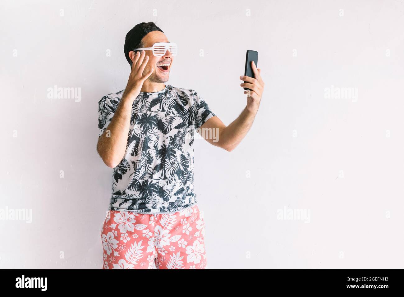 Jeune homme dans une chemise fleurie, casquette et lunettes en été, prenant  un selfie et une vidéoconférence avec son smartphone et boire un cocktail  avec une paille Photo Stock - Alamy