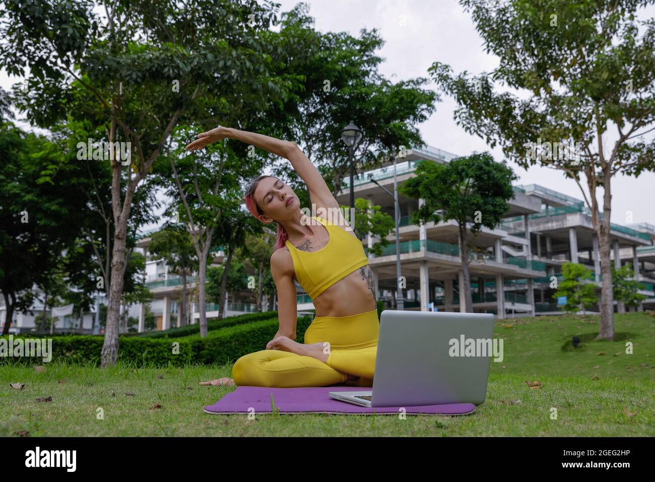 Jeune belle femme faisant du yoga dans le parc avec vidéo sur l'ordinateur portable. Regarder l'écran. Caucasien. Vêtements de sport colorés. Haute qualité Banque D'Images