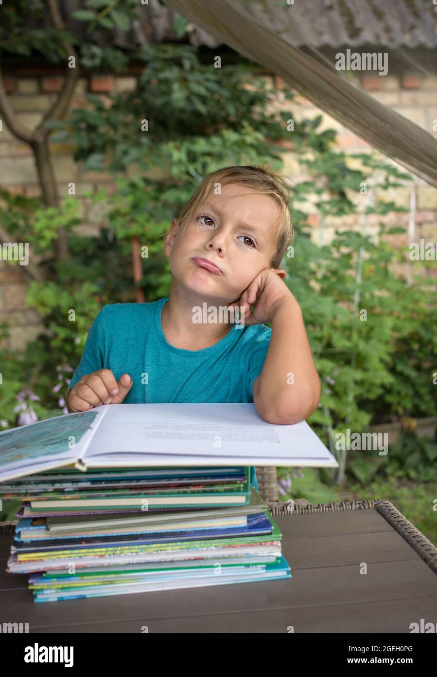 Garçon ennuyé de 5-6 ans assis à une table devant une pile de livres. Émotion du mécontentement, fatigue d'un préchoeur. Développement précoce, apprentissage Banque D'Images