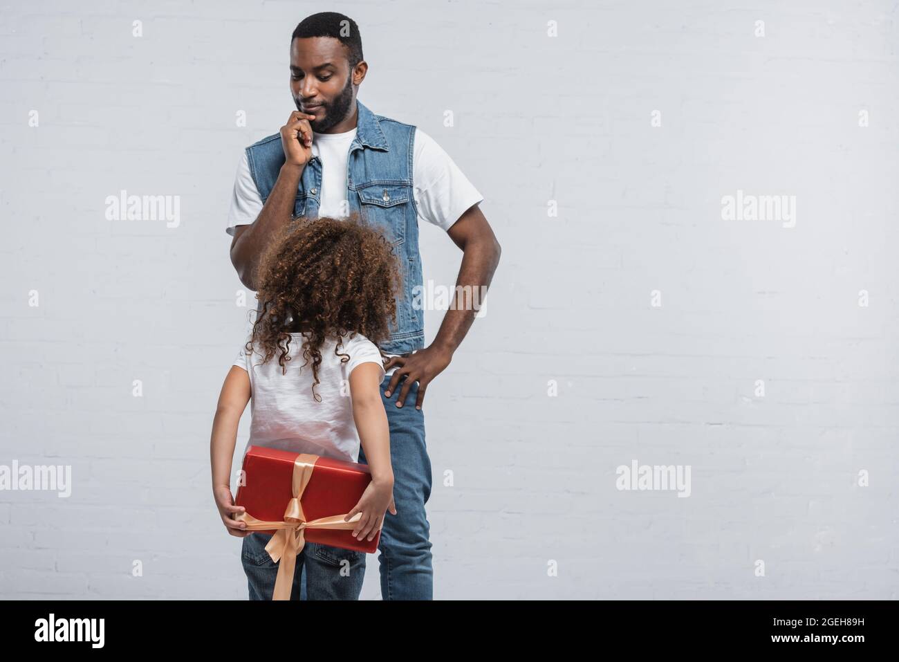 afro-américain sérieux debout avec la main sur la hanche près de la fille avec le présent derrière le dos sur le gris Banque D'Images