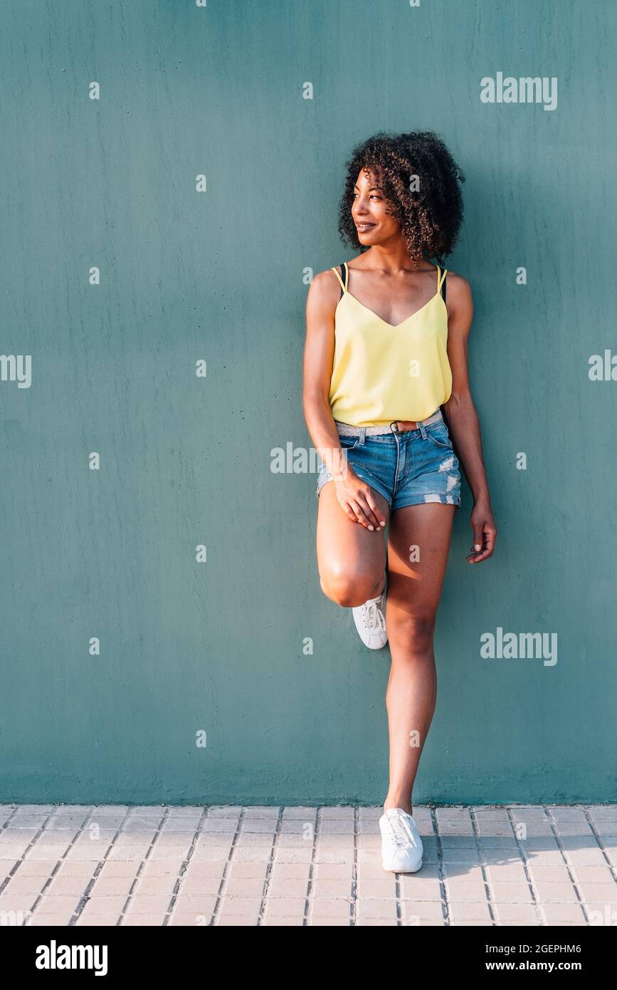 portrait vertical d'une femme africaine avec un fond de mur vert. Elle se penche sur le mur vert avec des vêtements d'été et souriant Banque D'Images