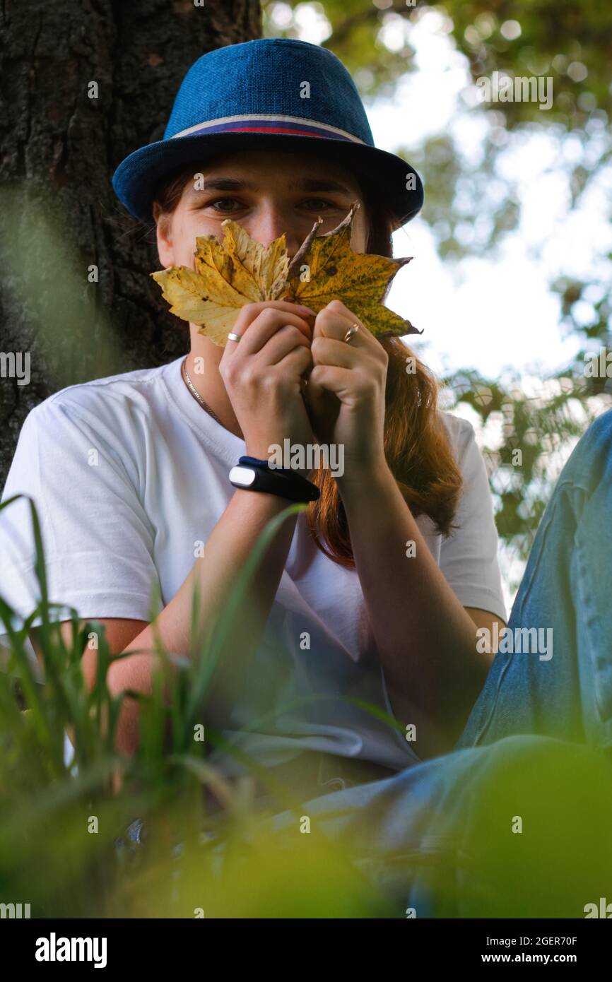 Portrait flou artistique d'une jeune femme drôle avec des cheveux bruns portant un chapeau à l'extérieur. Main femelle tenant des feuilles jaunes sèches et le visage couvert. Bonjour automne Banque D'Images