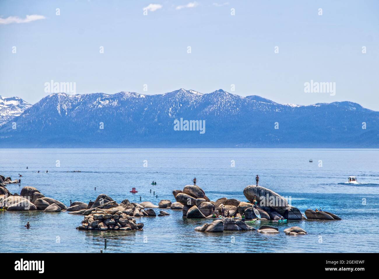 Touristes dans l'eau sur des paddleboards et escalade sur des rochers dans le lac Tahoe bleu avec des montagnes enneigées à distance Banque D'Images
